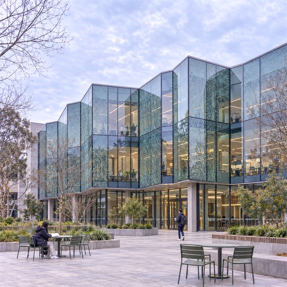 维多利亚大学 Footscray Park 校区:维多利亚大学 Footscray Park P 楼南庭院的冬季阴天示意图,灰绿折面玻璃带抽象蚀刻纹,一名学生阅读,另一名学生步行。